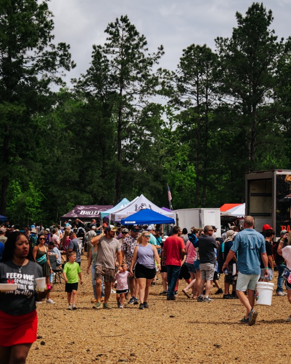 7th Annual Crawfish Critic Cookoff crowd flowing between cook tents under Conroe pines
